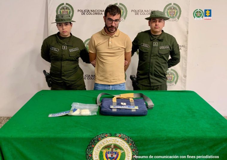 El capturado aparece detrás de una mesa, en la que están los elementos incautados. A cada lado suyo hay miembros de la Policía.