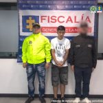 Se visualiza al capturado junto a un uniformado de la Policía Nacional e investigador del CTI. Detrás de ellos el banner que identifica a la Fiscalía General de la Nación.