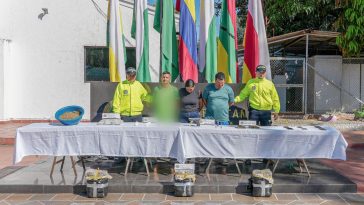 Los capturados se encuentran de frente y custodiados por dos uniformados de la Policía Nacional. En la foto se ve de fondo unas banderas, una mesa con el material incautado. En el piso se observa el estupefaciente incautado durante los registros de allanamientos.