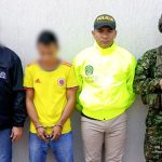 El capturado se encuentra de frente y tiene una camiseta de la selección Colombia. Se encuentra custodiado por un servidor del Cuerpo Técnico de Investigación CTI, un uniformado de la Policía y Ejército Nacional.  