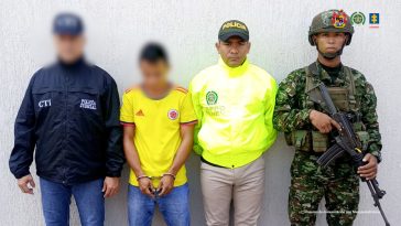 El capturado se encuentra de frente y tiene una camiseta de la selección Colombia. Se encuentra custodiado por un servidor del Cuerpo Técnico de Investigación CTI, un uniformado de la Policía y Ejército Nacional.  