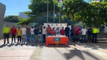 En la fotografía aparecen personas de pie, con los brazos atrás, esposados, custodiados por dos uniformados de la Policía Nacional. Detrás de ellos hay un pendón de esa institución y delante de ellos una mesa con los elementos incautados. 