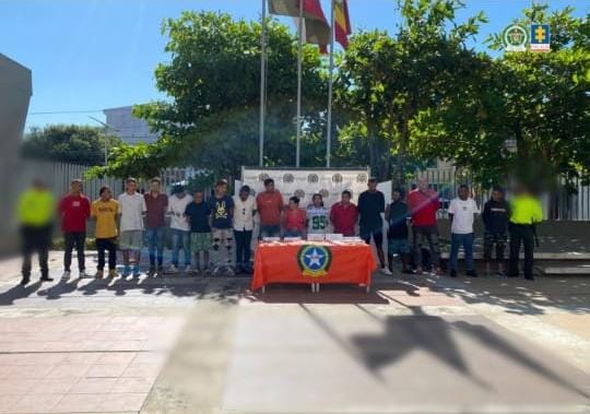 En la fotografía aparecen personas de pie, con los brazos atrás, esposados, custodiados por dos uniformados de la Policía Nacional. Detrás de ellos hay un pendón de esa institución y delante de ellos una mesa con los elementos incautados. 