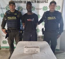 En la fotografía se aprecia de pie y con los brazos atrás a Carlos Andrés Cuero Vallecilla. Viste con camiseta y pantalón de color negro. A sus extremos hay uniformados de la Policía Nacional. delante de ellos hay una superficie con mantel blanco y sobre ella un letrero que dice: ‘Captura por orden judicial’. Detrás de ellos hay un pendón institucional de la Policía.