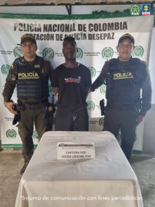 En la fotografía se aprecia de pie y con los brazos atrás a Carlos Andrés Cuero Vallecilla. Viste con camiseta y pantalón de color negro. A sus extremos hay uniformados de la Policía Nacional. delante de ellos hay una superficie con mantel blanco y sobre ella un letrero que dice: ‘Captura por orden judicial’. Detrás de ellos hay un pendón institucional de la Policía.