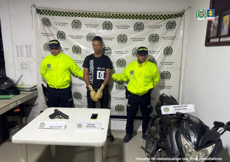 El capturado aparece en el centro de la imagen, con las manos esposadas. Frente a él se ve un arma y una motocicleta. Dos miembros de la Policía rodean al hombre.