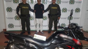 El capturado se encuentra de frente y  custodiado por dos uniformados de la Policía Nacional. En la foto se ve de fondo un backing de la Policía Nacional y la motocicleta que se había hurtado.