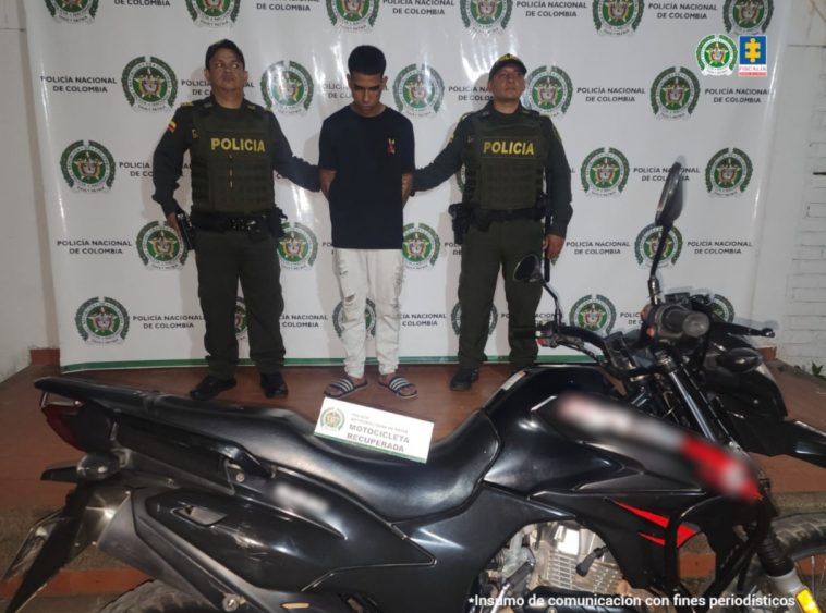 El capturado se encuentra de frente y  custodiado por dos uniformados de la Policía Nacional. En la foto se ve de fondo un backing de la Policía Nacional y la motocicleta que se había hurtado.