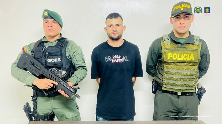 En la fotografía se aprecia de pie y con los brazos atrás a Jhon Fredy Ramírez Bedoya, alias Barbado. Usa barba y viste camiseta de manga corta de color negro, con estampado a la altura del pecho. A sus extremos hay personal de la Policía Nacional. Delante de ellos hay una superficie y sobre ella hay un letrero que dice: ‘Captura por orden judicial’.