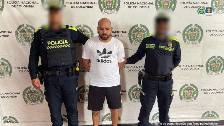 El capturado aparece de pie, en medio de dos miembros de la Policía. Está esposado con las manos atrás. A sus espaldas puede verse un banner de la Policía.