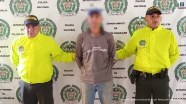 Se observa al capturado de frente y un efecto hace su rostro borroso.  A cada lado lo custodia un funcionario de la Policía Nacional. Atrás, u pendón de Policía Nacional