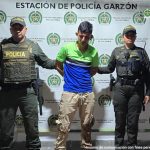 El capturado se encuentra de frente y custodiado por dos uniformados de la Policía Nacional. En la foto se ve de fondo un backing de la Policía Nacional.