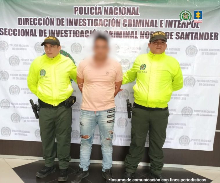 Se visualiza al capturado junto a dos uniformados de la Policía Nacional. Detrás el banner institucional que identifica a la Dirección de Investigación de la Policía Nacional en Norte de Santander 