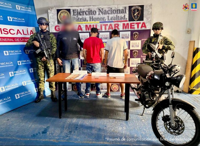 Capturados de espaldas custodiados por investigador del CTI de la Fiscalía y militares del Ejército Nacional. Delante mesa con material incautado . Detrás banner de la Fiscalía y del Gaula Militar.