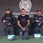   En la imagen se observa al capturado junto a dos  uniformados de la Policía Nacional.  Frente a ellos una mesa con los elementos incautados. Detrás el banner que identifica a la Policía Nacional.