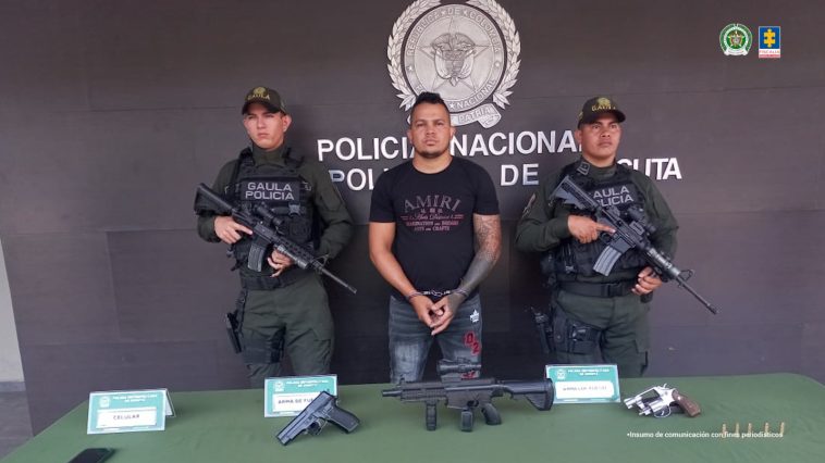   En la imagen se observa al capturado junto a dos  uniformados de la Policía Nacional.  Frente a ellos una mesa con los elementos incautados. Detrás el banner que identifica a la Policía Nacional.