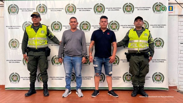 Los capturados se encuentran de frente y  custodiados por dos uniformados de la Policía Nacional. En la foto se ve de fondo un backing de la Policía Nacional.