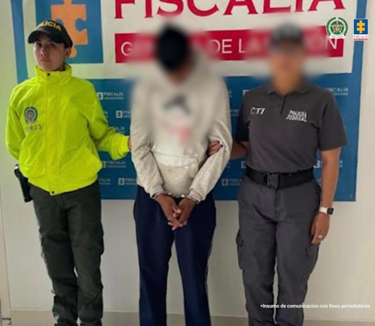 El capturado aparece esposado, en medio de una servidora de la Fiscalía y una de la Policía. Al fondo se ve un banner de la Fiscalía