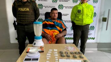 En la fotografía se aprecia sentado en silla de ruedas Juan Gildardo Chávez Quintero. Viste con una ruana de franjas de colores naranja, negro y azul. A sus extremos hay uniformados de la policía Nacional. Delante de ellos hay una superficie y sobre ella hay bolsas plásticas transparentes, pequeñas. Unas tienen contenido blano y otras contenido vegetal verdoso; asimismo, una gramera y una licuadora. Detrás de ellos hay un pendón institucional.