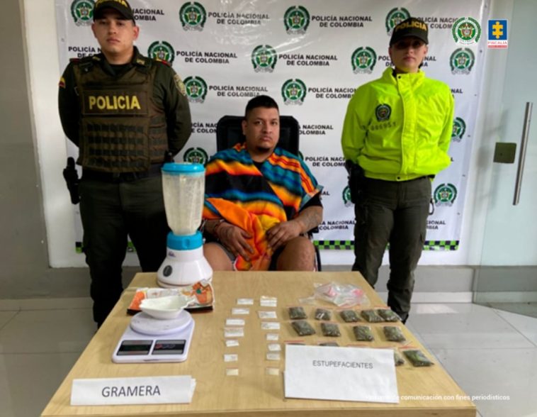 En la fotografía se aprecia sentado en silla de ruedas Juan Gildardo Chávez Quintero. Viste con una ruana de franjas de colores naranja, negro y azul. A sus extremos hay uniformados de la policía Nacional. Delante de ellos hay una superficie y sobre ella hay bolsas plásticas transparentes, pequeñas. Unas tienen contenido blano y otras contenido vegetal verdoso; asimismo, una gramera y una licuadora. Detrás de ellos hay un pendón institucional.