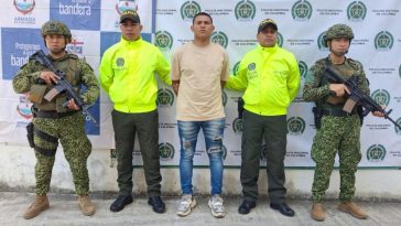 El capturado aparece en el centro de la imagen, rodeado de dos miembros de la Policía y dos miembros del Ejército. Al fondo puede verse un banner de la Policía y uno del Ejército.