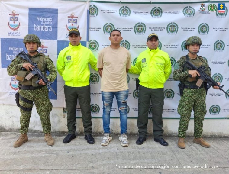 El capturado aparece en el centro de la imagen, rodeado de dos miembros de la Policía y dos miembros del Ejército. Al fondo puede verse un banner de la Policía y uno del Ejército.