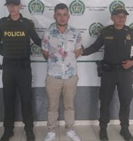 En la imagen se ve una persona detenida bajo custodia de dos integrantes de la Policía Nacional. Detrás suyo un backing institucional.