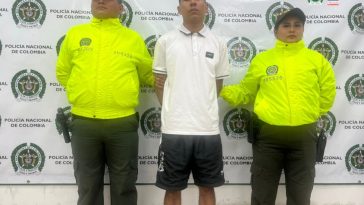 Un hombre aparece en el centro de la imagen, rodeado de dos miembros de la Policía. Hay un banner de la Policía en el fondo.