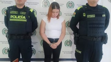 En la fotografía se aprecia de pie, con los brazos adelante y esposada a María Corona Arango Grajales. Viste con blusa blanca de manga en el hombre y pantalón negro. Usa cabello de color naranja y raiz oscura. A sus extremos hay uniformados de la Policía Nacional. Delante de ellos hay un superficie de color madera y sobre ella un letrero que dice: ‘Orden judicial’. Detrás de ellos hay un pendón institucional de la Policía.