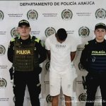 El hombre aparece en el centro de la imagen, con la cabeza agachada y las manos esposadas en la espalda. Lo rodean miembros de la Policía y al fondo hay un backing de la misma institución.