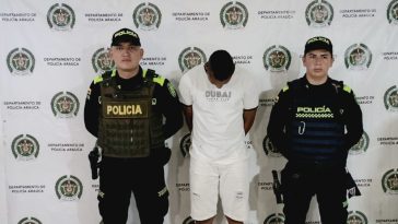 El hombre aparece en el centro de la imagen, con la cabeza agachada y las manos esposadas en la espalda. Lo rodean miembros de la Policía y al fondo hay un backing de la misma institución.