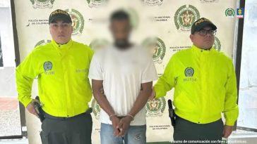 En la fotografía está un hombre de pie, con el rostro difuminado, vestido con camiseta blanca, se encuentra con las manos adelante esposadas y custodiado por dos uniformados de la Policía Nacional.