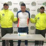 En la foto aparece un hombre trigueño, de pie, con la cabeza agachada, brazos atrás esposado, vestido con una camiseta blanca, azul y naranja y un jean azul. Se encuentra custodiado por dos uniformados de la Policía Nacional. 