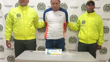 En la foto aparece un hombre trigueño, de pie, con la cabeza agachada, brazos atrás esposado, vestido con una camiseta blanca, azul y naranja y un jean azul. Se encuentra custodiado por dos uniformados de la Policía Nacional. 