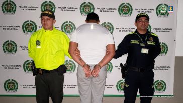 capturado de espaldas custodiado por dos policías. Detrás banner de la Policía Nacional.