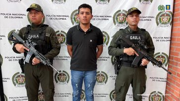 capturado de frente custodiado por dos policías. Detrás banner de la Policía Nacional.