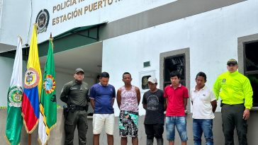 se observa a los capturados custodiados por dos uniformados de la Policía Nacional, están ubicados al frente a la estación de Policía de Puerto Nariño y a la derecha de los capturados se observa las banderas de Colombia y Amazonas