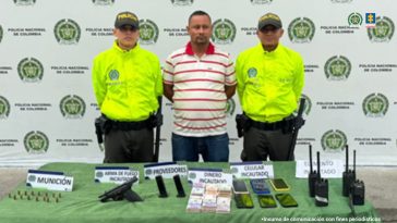 En la fotografía se aprecia de pie y con los brazos atrás a León Jaime Urrego Restrepo. Viste con camisa de color blanco con franjas rojas horizontales, manca corta y cuello; jean azul. Usa barba tipo candado. A sus extremos hay uniformados de la Policía Nacional. Delante de ellos hay una superficie de color verde y sobre ella hay un arma de fuego, munición, proveedores, dinero en efectivo, varios celularesny dos radios de comunicaciones. Detrás de ellos hay un pendón institucional de la Policía