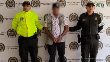 En la imagen se ve una persona detenida bajo custodia de dos integrantes de la Policía Nacional. Detrás suyo un backing institucional.