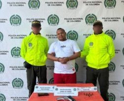 En la imagen está un hombre de estatura mediana, contextura gruesa, moreno, de cabellos cortos negros crespos, con las manos adelante esposadas, vestido con una camiseta blanca y un pantalón rojo, custodiado por dos uniformados de la Policía Nacional. 