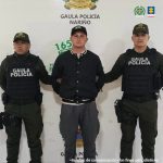 El capturado se encuentra de frente y custodiado por dos uniformados del Gaula de la Policía Nacional. En la foto se ve de fondo un backing de la Policía.