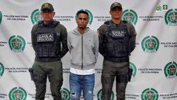 El hombre capturado aparece de pie, con las manos atrás, en medio de dos miembros de la Policía. Al fondo puede verse un banner de la misma institución.