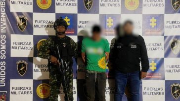 capturado de frente custodiado por investigador del CTI de la Fiscalía y un soldado. Detrás banner del grupo Gaula Militar.