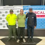 Se visualiza al capturado junto a un uniformado de la Policía Nacional y un investigador del CTI. Detrás de ellos los banners que identifican a la Policía Nacional y a la Fiscalía General de la Nación.