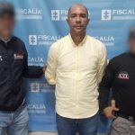 En la fotografía está un  hombre moreno de pie, semi calvo, con los brazos atrás, vestido con una camisa manga larga amarilla y un jean azul, custodiado por dos uniformados del CTI. Detrás de ellos hay un pendón de la Fiscalía. 