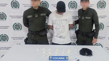 En la fotografía aparece un hombre de pie, con gorra negra, tiene la cabeza agachada y los brazos atrás esposado. Se encuentra custodiado por dos uniformados de la Policía Nacional. Delante de ellos está la droga y la peluca incautada.