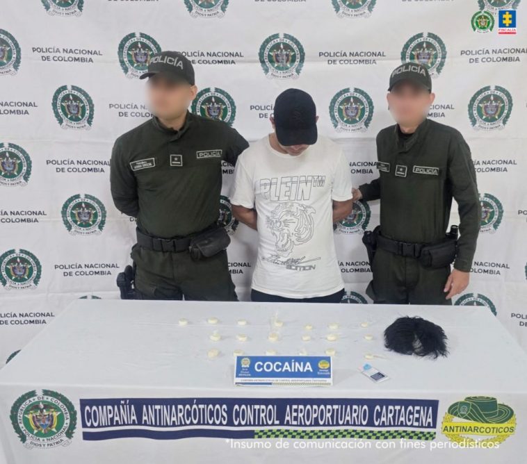 En la fotografía aparece un hombre de pie, con gorra negra, tiene la cabeza agachada y los brazos atrás esposado. Se encuentra custodiado por dos uniformados de la Policía Nacional. Delante de ellos está la droga y la peluca incautada.