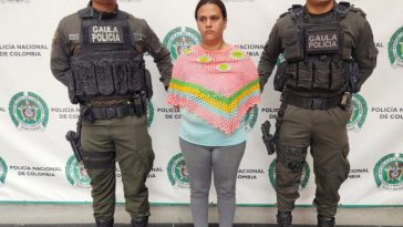 En la imagen se ve una mujer detenida bajo custodia de dos uniformados de la Policía Nacional. Detrás suyo un backing institucional.